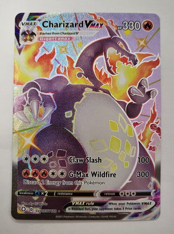 Holografische Pokémon-VMAX-Karte von Charizard VMAX mit 330 KP. Die Karte zeigt Glurak in seiner Gigadynamax-Form, umgeben von Flammen, mit einem dunklen, schimmernden Design. Enthält die Attacken ‚Claw Slash‘ und ‚G-Max Wildfire‘. Als VMAX-Karte gibt sie dem Gegner drei Preiskarten, wenn sie besiegt wird.
