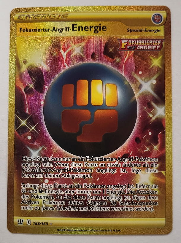 Goldene Spezial-Energiekarte ‚Fokussierter-Angriff-Energie‘ aus dem Pokémon-Sammelkartenspiel. Die Karte zeigt ein leuchtendes, goldenes Faust-Symbol vor einem dunklen, funkelnden Hintergrund. Sie liefert Kampf- und Finsternis-Energie und verstärkt die Attacken eines angelegten Pokémon um 20 Schadenspunkte.