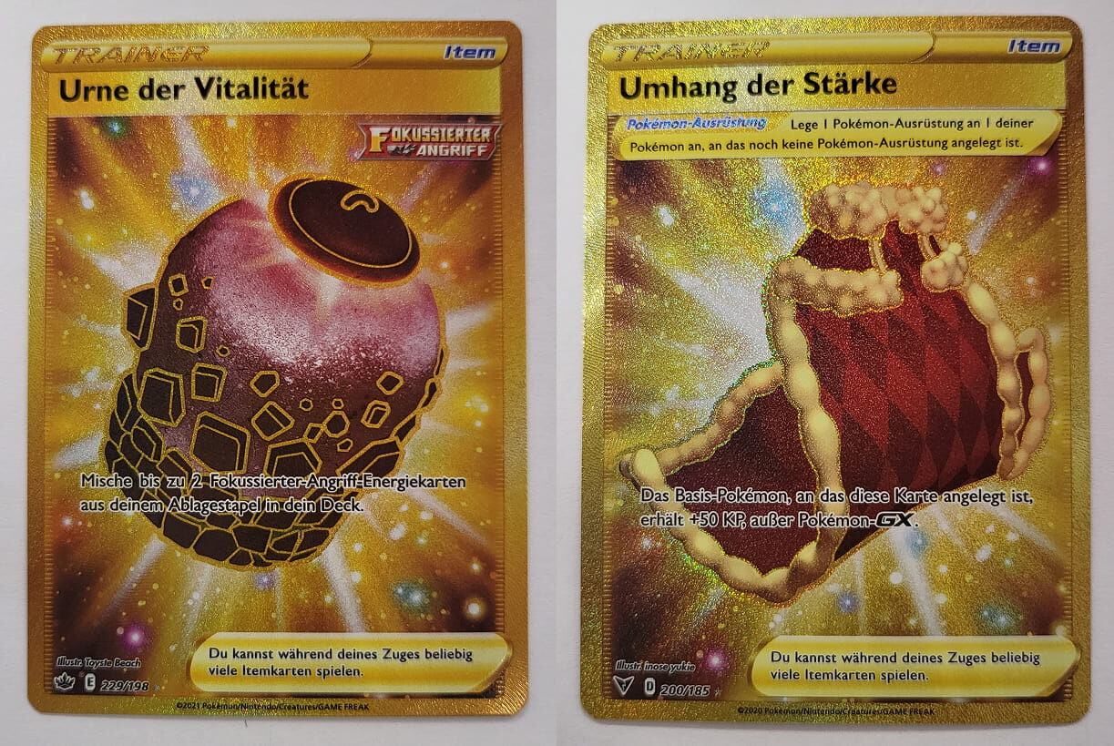 Zwei goldene Pokémon-Item-Karten nebeneinander: Links ‚Urne der Vitalität‘ mit einem leuchtenden Urnen-Design, die es ermöglicht, bis zu zwei Fokussierter-Angriff-Energiekarten ins Deck zu mischen. Rechts ‚Umhang der Stärke‘ mit einem roten, pelzbesetzten Umhang, der dem angelegten Basis-Pokémon 50 zusätzliche KP verleiht. Beide Karten haben ein holografisches Gold-Finish.