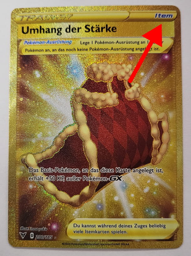 Goldene Pokémon-Trainerkarte ‚Umhang der Stärke‘ mit leuchtendem Effekt. Eine rote Pfeilmarkierung hebt das ‚Item‘-Label in der oberen rechten Ecke hervor. Die Karte verleiht einem Basis-Pokémon 50 zusätzliche KP.