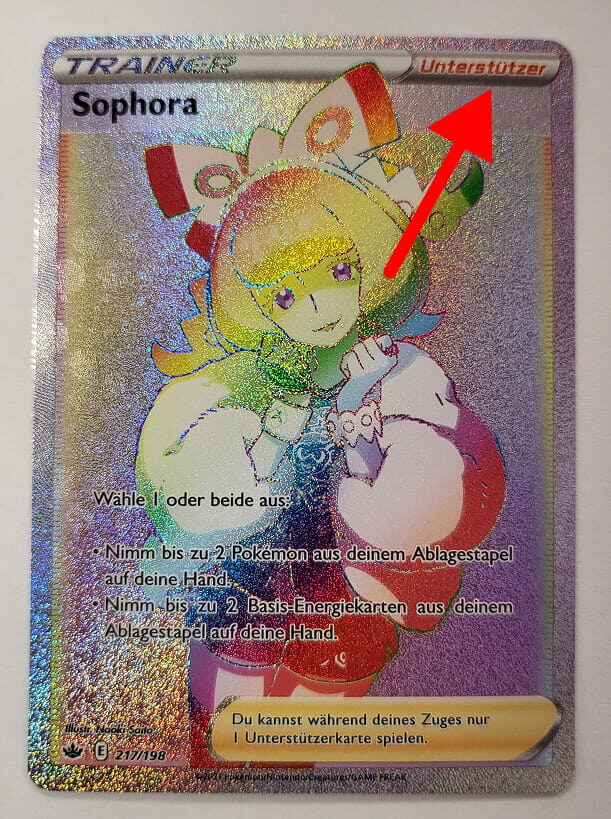 Holografische Pokémon-Trainerkarte ‚Sophora‘ mit einem bunten, regenbogenfarbenen Schimmer. Eine rote Pfeilmarkierung hebt die Kategorie ‚Unterstützer‘ in der oberen rechten Ecke hervor.